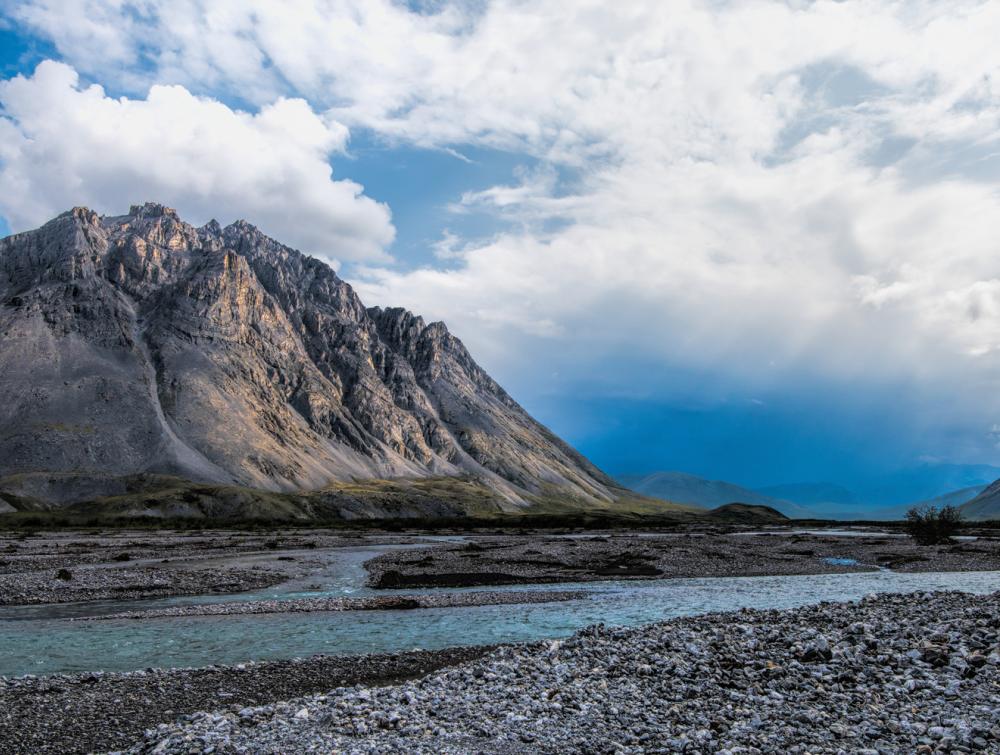 Cloudy skies above mountains and a river bordered by coarse rocky beaches