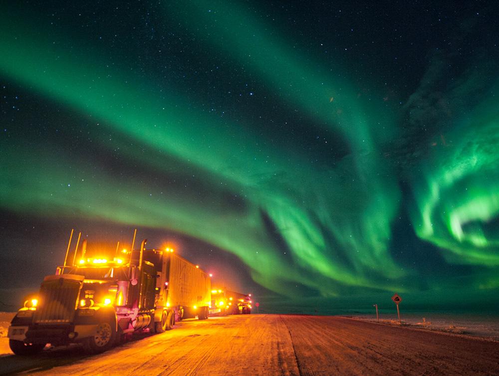 A truck drives past oil & gas production at night with its lights on. A green aurora is shown in a swirling pattern in the night sky.