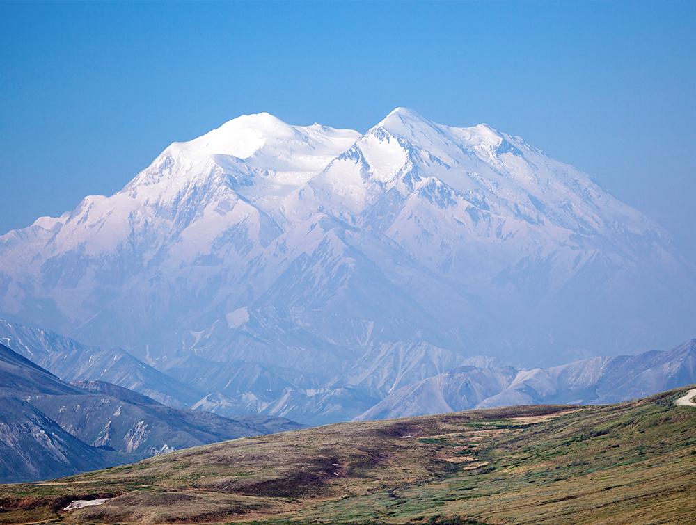 A mountain fills a hazy image with snowy peaks and a blue sky with a basic landscape in the foreground.