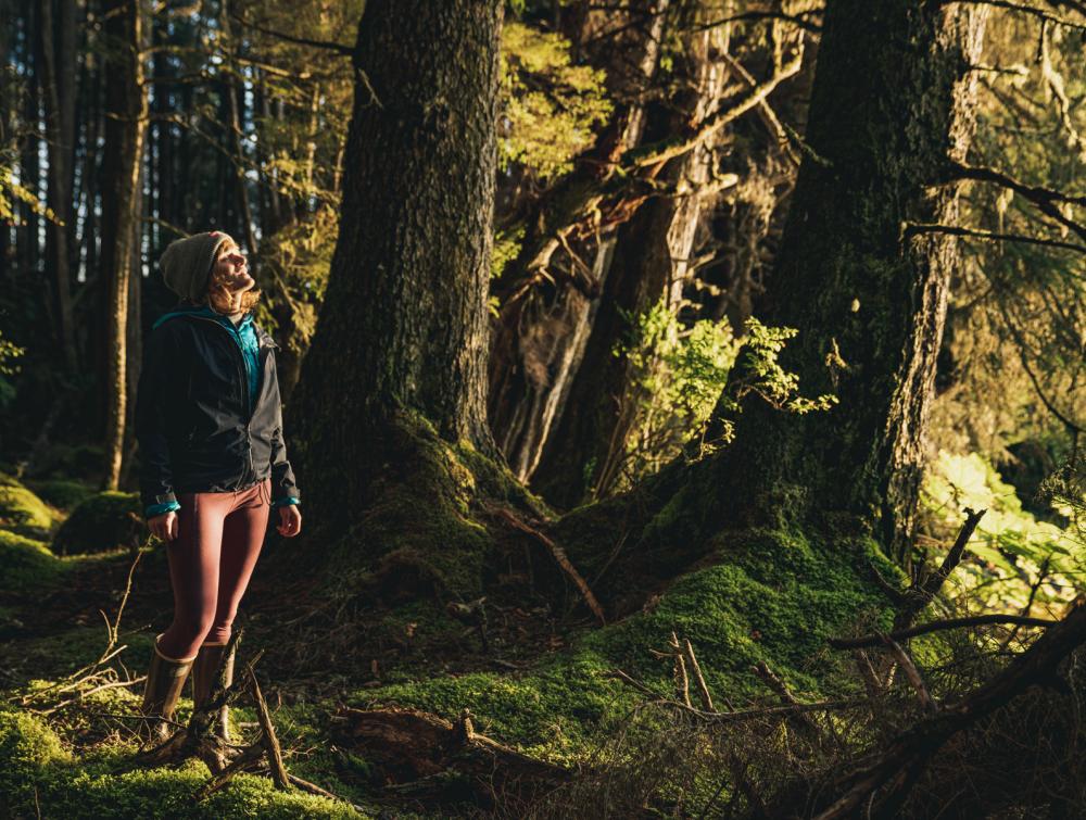 Person with hat and jacket looking to the right of the frame toward a source of light filtering through mossy trees