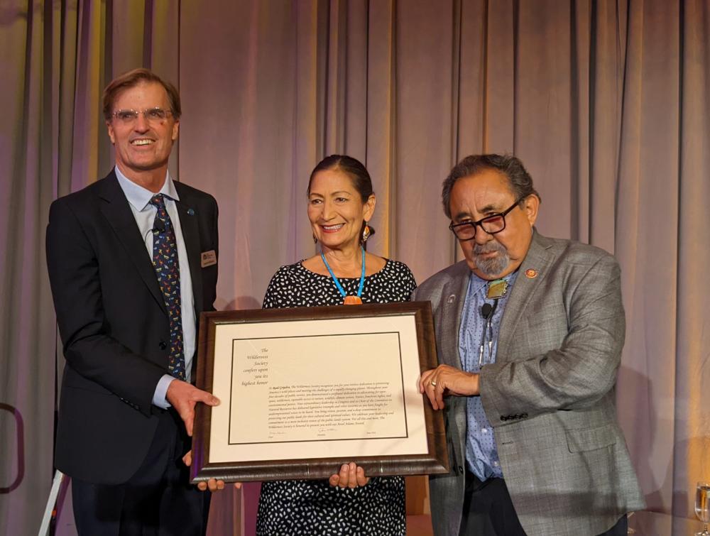 Rep. Raul Grijalva, Interior Secretary Deb Haaland and Wilderness Society President Jamie Williams standing next to each other in front of a curtain, holding a large framed document