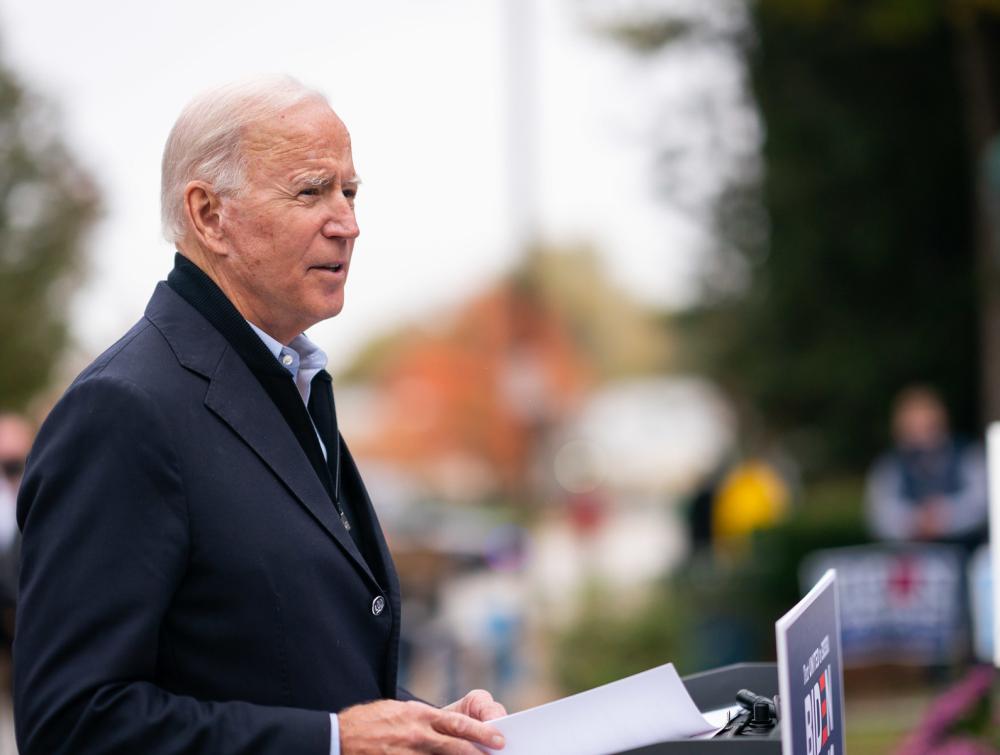 Joe Biden looking to the right and holding papers with trees and buildings in the background