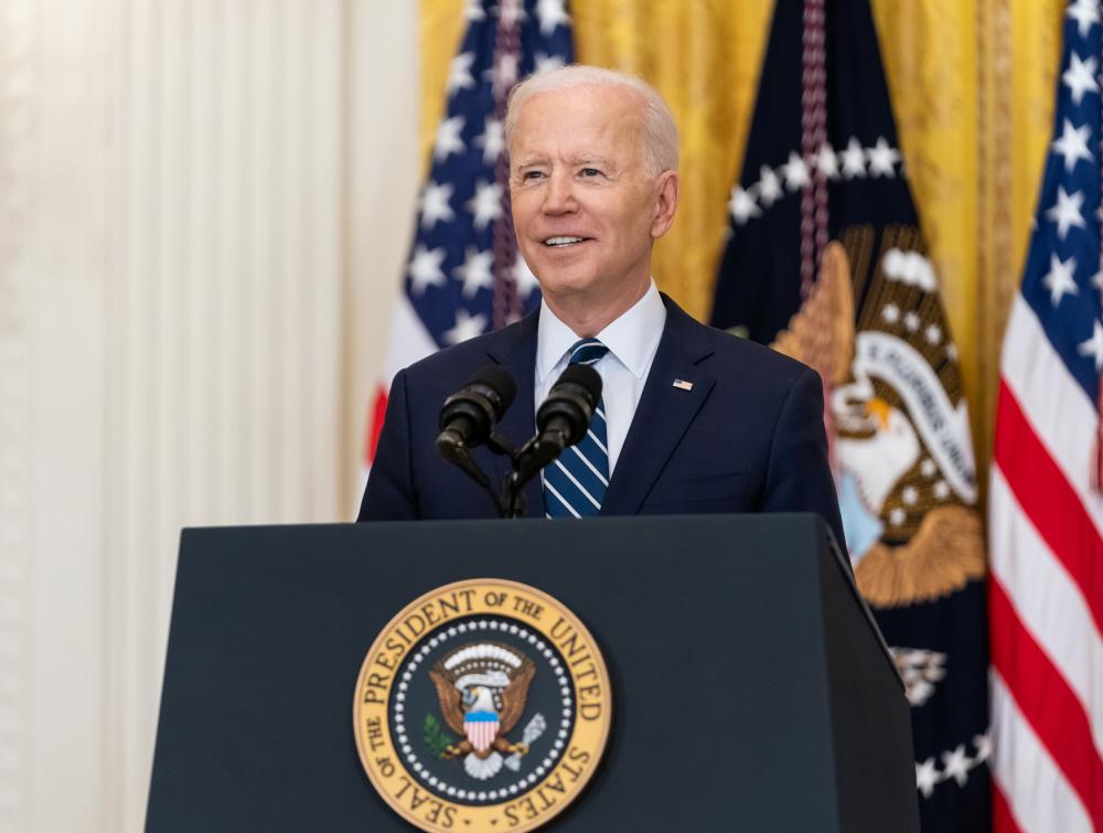 President Joe Biden facing forward, standing behind a lectern with the presidential seal on it and in front of American flags