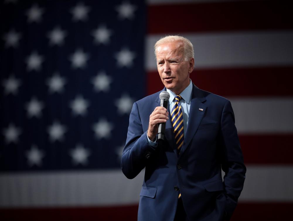 President Joe Biden holding a microphone and standing in front of a large American flag backdrop