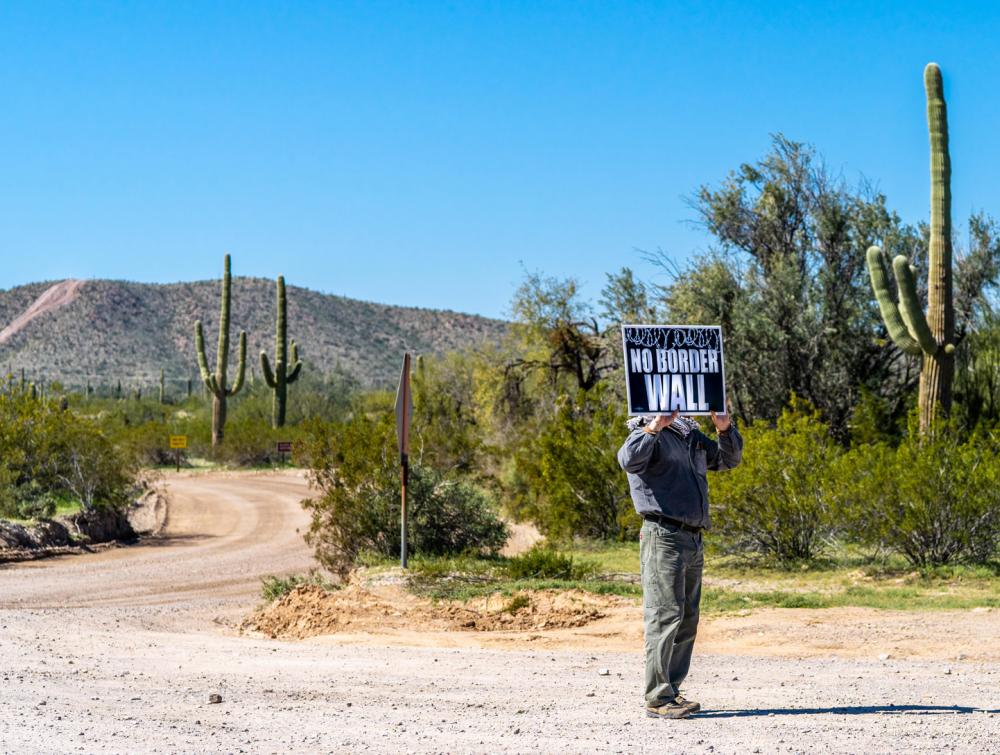 Person standing with sign that reads "No Border Wall" underneath blue sky with green cactus and other plants behind them