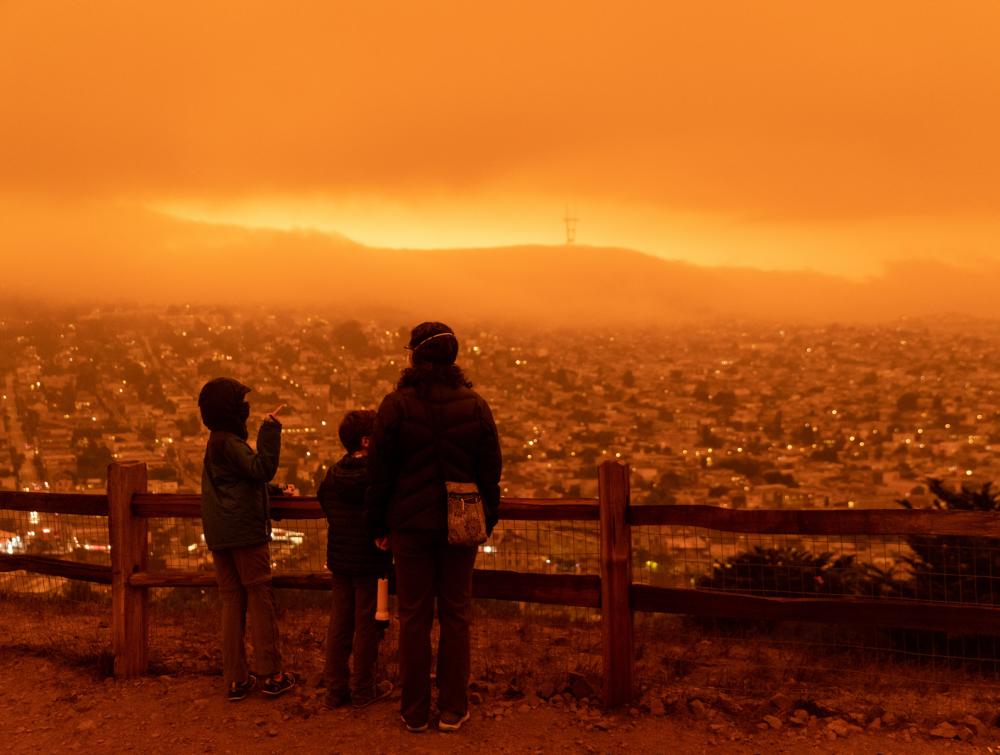 Three people standing in front of a fence looking out over a cityscape with an all-encompassing hazy orange light overwhelming the scene