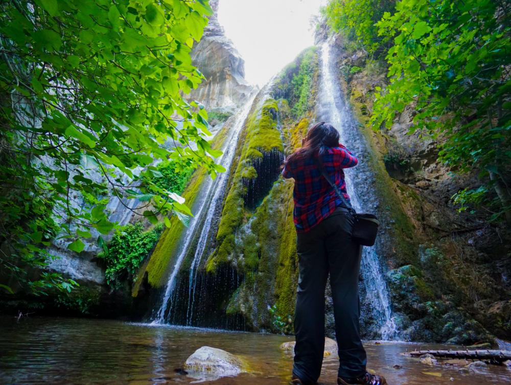 Person pictured from rear looking up and taking picture of waterfall surrounded by greenery