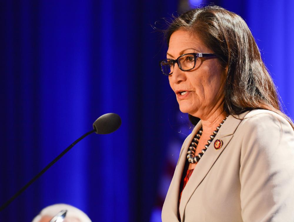 Woman with glasses speaking into microphone in front of blue curtain