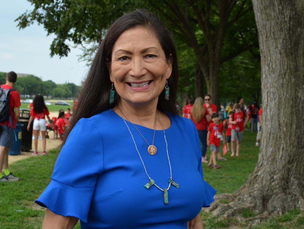 Person with long dark hair, wearing blue dress and facing camera with green grass, trees, and a crowd in the background