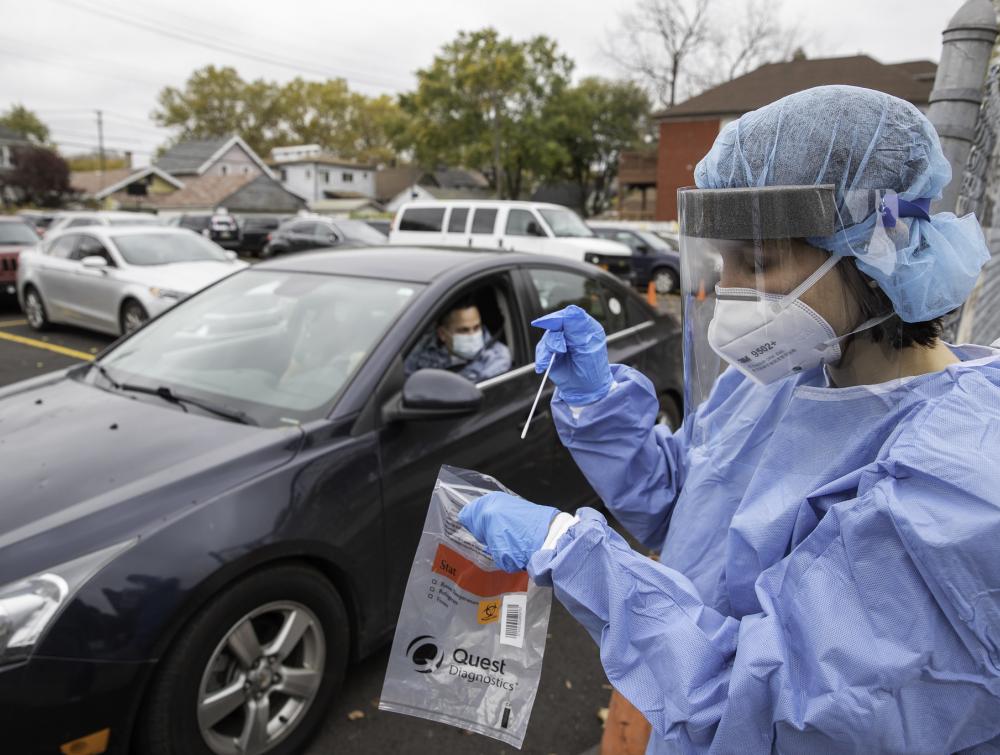 Medical worker wearing personal protective equipment and holding COVID nasal swab and specimen bag. Driver in car is in the background.