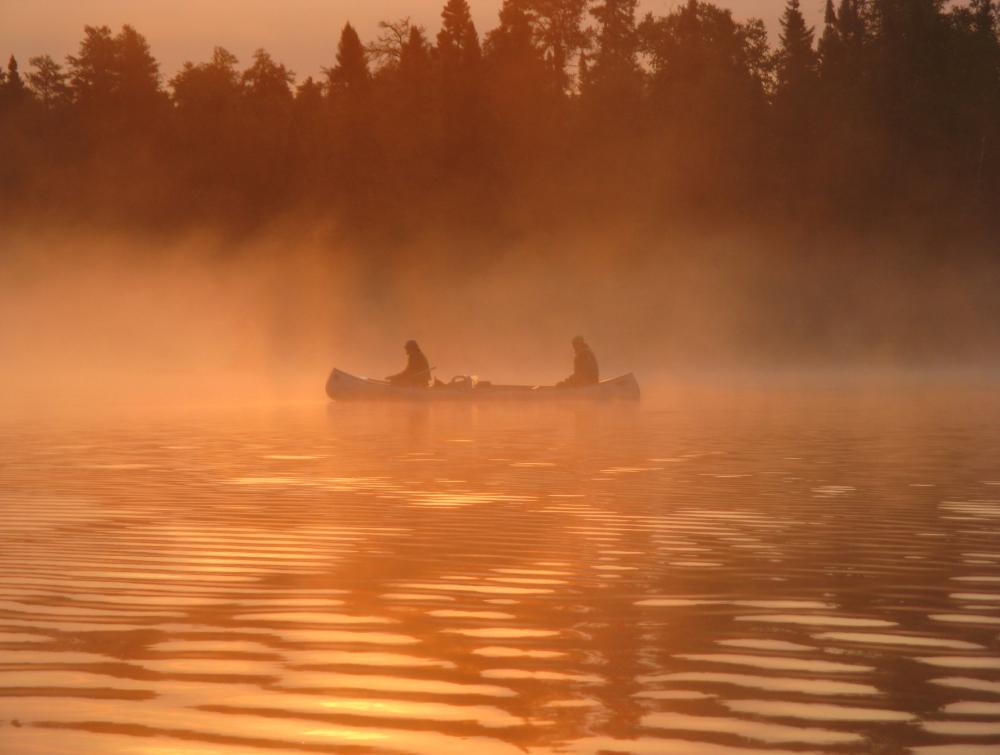 Two people in a canoe on a lake, partly obscured by haze in an orange partly sunlit scene