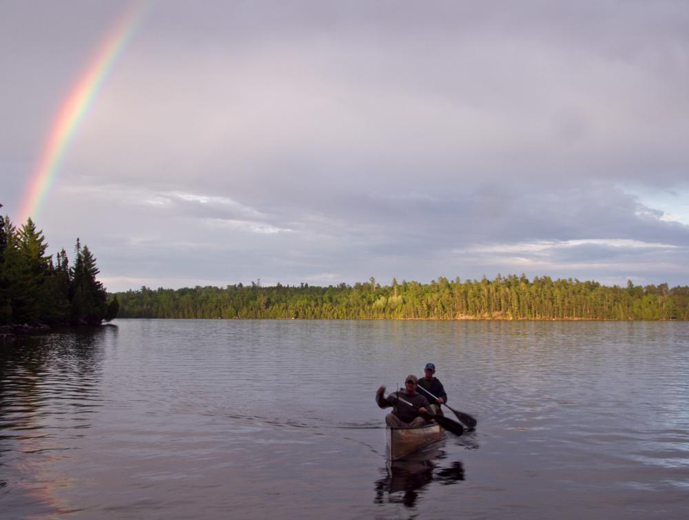Two people in a canoe paddling toward foreground in a lake with a rainbow in the upper left corner, Boundary Waters Canoe Area Wilderness, Minnesota