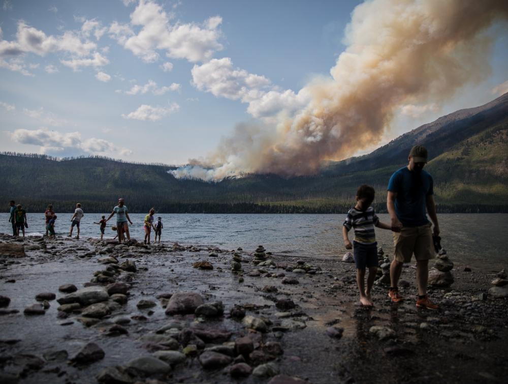 People walking along the edge of a lake in the foreground with forested landscape and a tall column of smoke visible in the distance