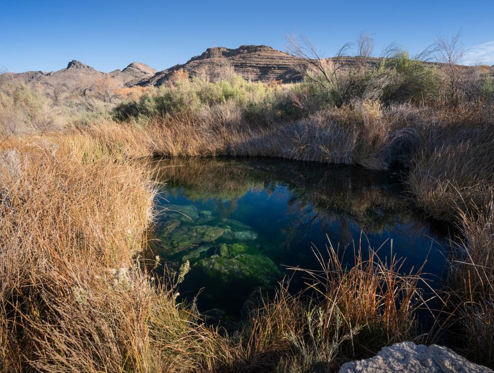 Brilliant blue body of water surrounded by desert grasses with rock formations in the background