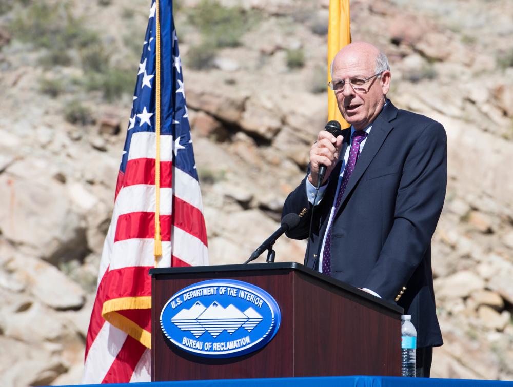 Man standing at lectern with microphone in his hand and an American flag in the background