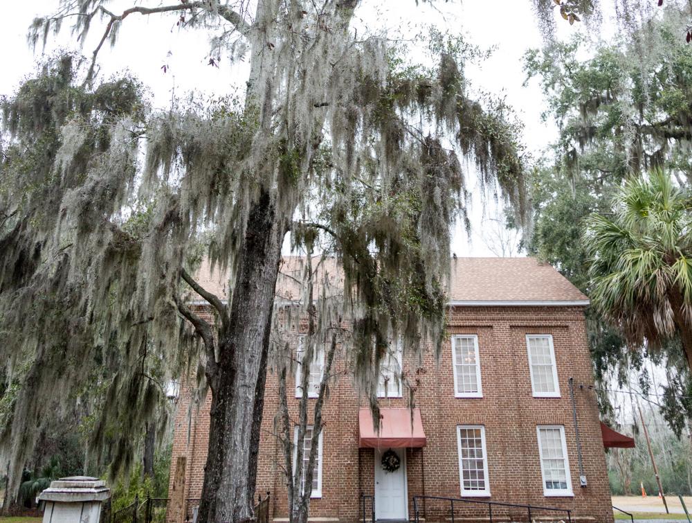 Brick building with many windows and small reddish awning atop front door. Spanish moss is visible hanging from tree branches in foreground.