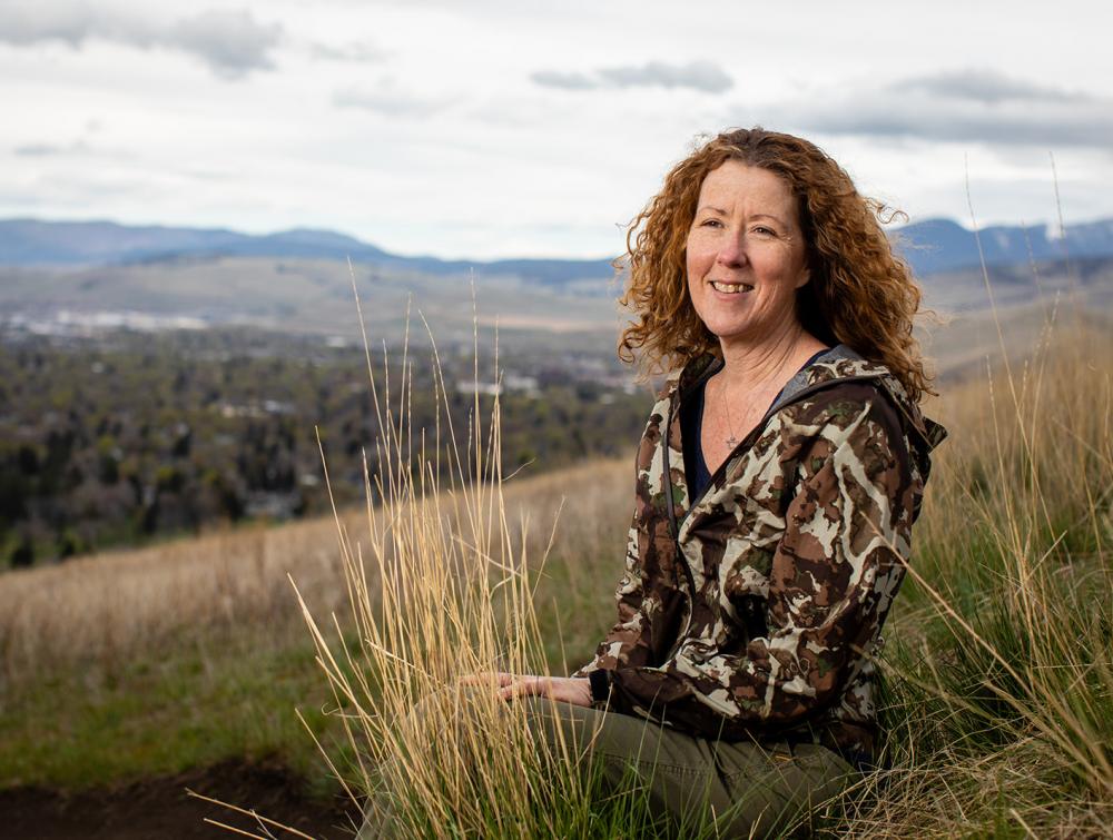 Woman sitting on grass hillside and looking into the middle distance toward the viewer, with rolling landscape and cloudy skies behind her