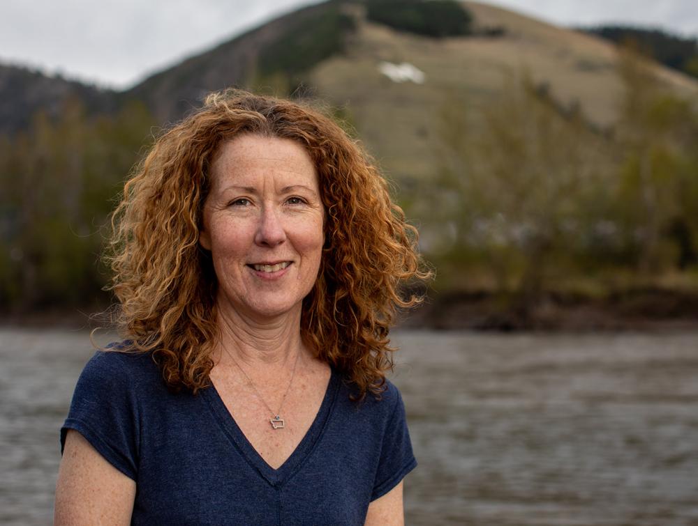 Person with long curly hair standing in front of river and mountainous landscape 