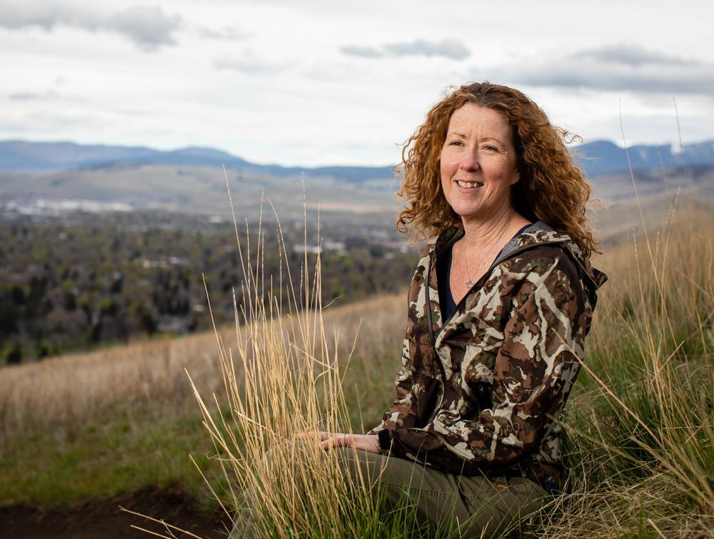 Person with long curly hair standing in front of river and mountainous landscape 