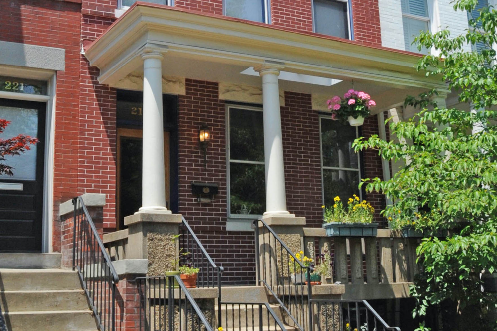 red brick two story home with white columns on the porch. There is a wrought iron stairwell on the left side of the stairs.