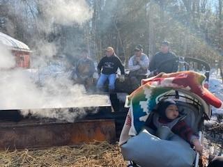 Many stand in front of a pot where sap boils, a child in a stroller is in the foreground.