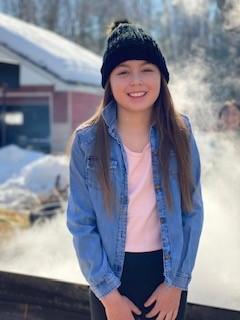 A child smiles in front of the sap pan boiling behind them.