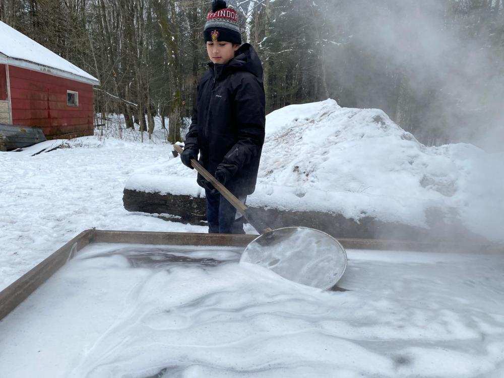 A young man dressed warmly skims off foam from the top of the sap boil. 