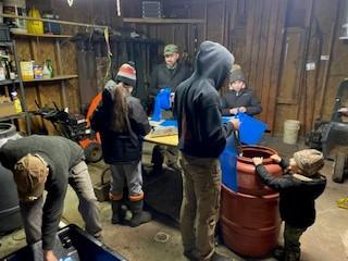 A family, wearing clothing for cold weather, prepares sap collection bags inside an enclosed area. 
