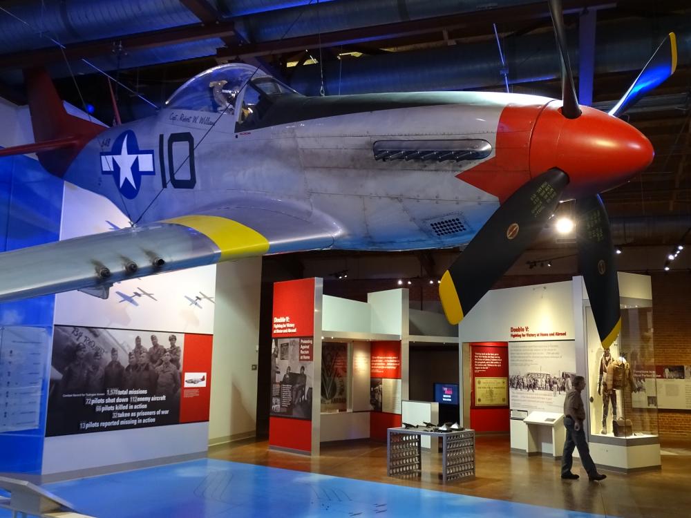Old warplane hanging from ceiling inside a museum, with pictures of pilots on the walls of the museum