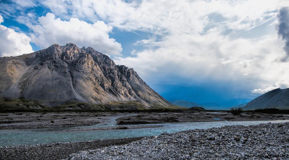 Cloudy skies above mountains and a river bordered by coarse rocky beaches
