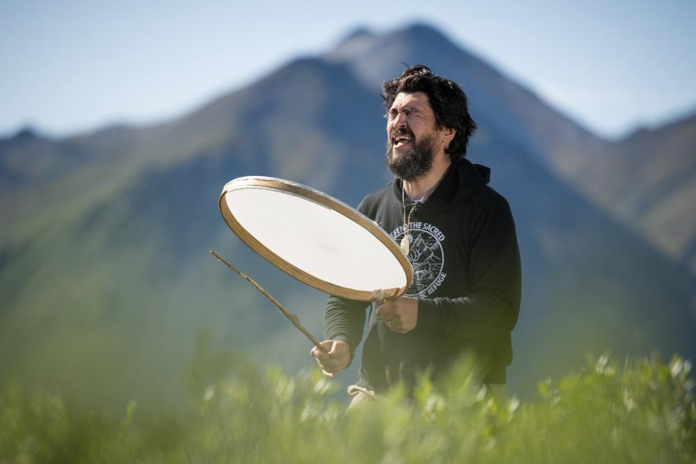 Bearded man using a stick to beat the face of a large, shallow drum with mountains in the background