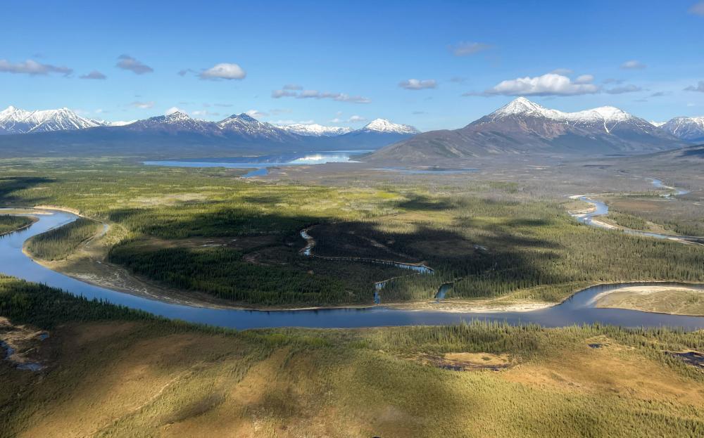 An aerial view of a winding river flowing through a vast wilderness valley, with snow-capped mountains rising in the distance under a clear blue sky.