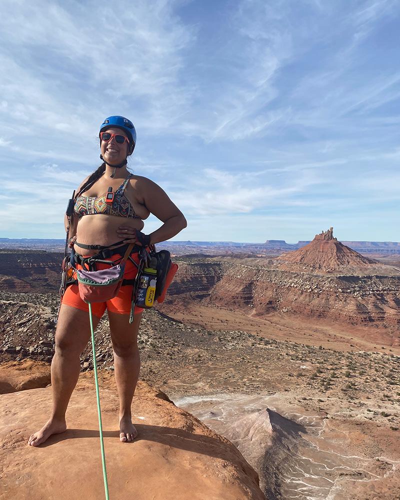 Bennett is wearing orange shorts and a patterned sports bra with a harness tied into a green rope. She is standing on top of a red sandstone rock with the North Six Shooter formation in the background 