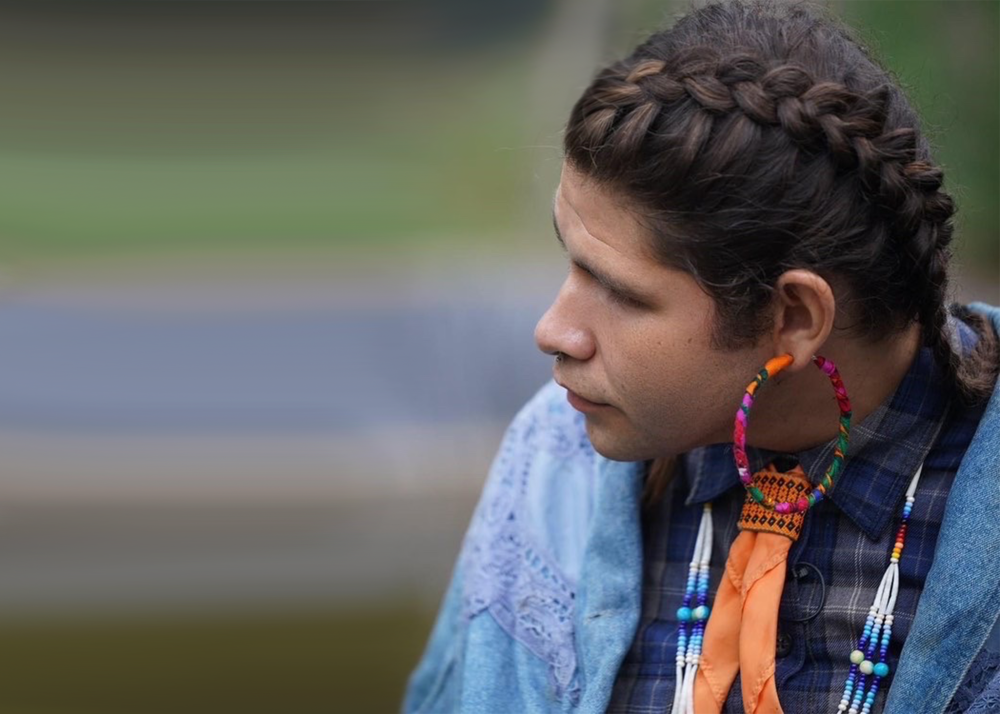 A two spirit person wears their hair in braids and looks to the left