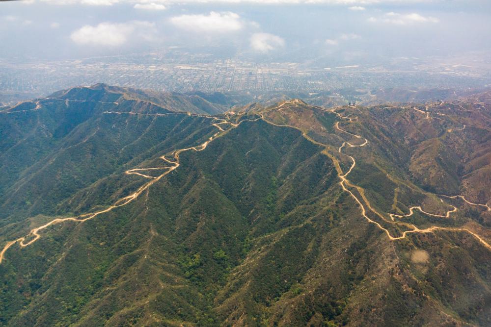 Aerial view of dirt path winding through small greenery-draped mountain range with city visible in the distance