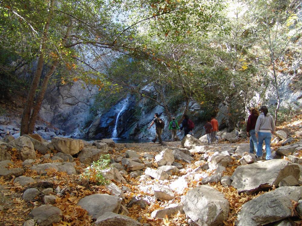 Group of people seen from rear hiking across rocks and fallen leaves toward waterfall in San Gabriel Mountains National Monument, California