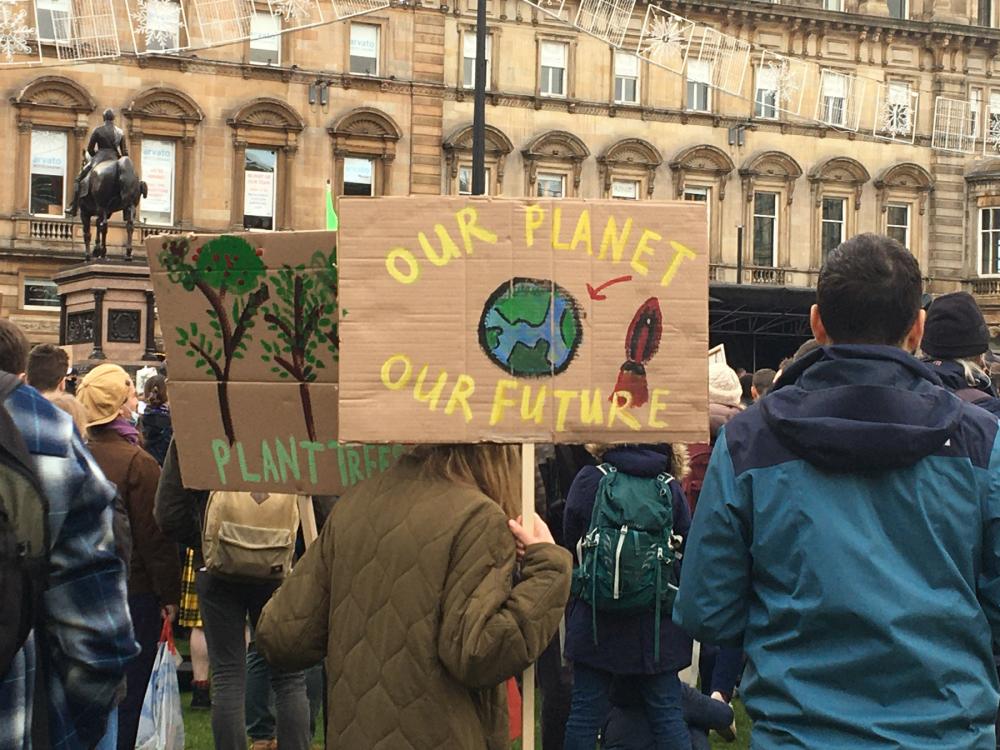 Crowd of people wearing coats and marching in front of a large building. Visible sign reads "OUR PLANET OUR FUTURE"