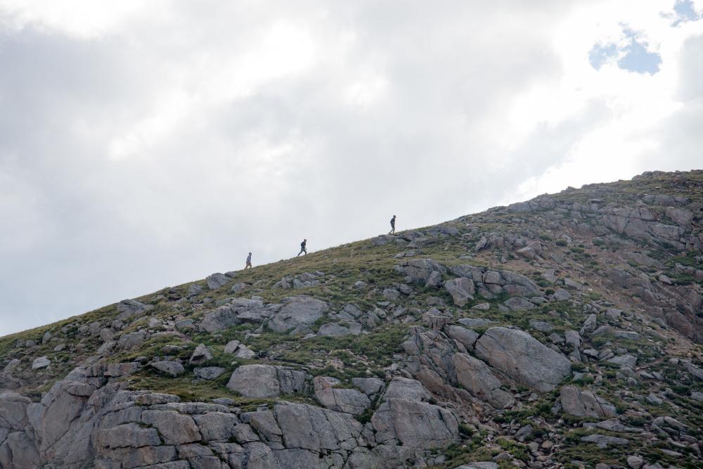 Three hikers seen from a distance, walking up a rock-strewn hillside under a cloudy sky