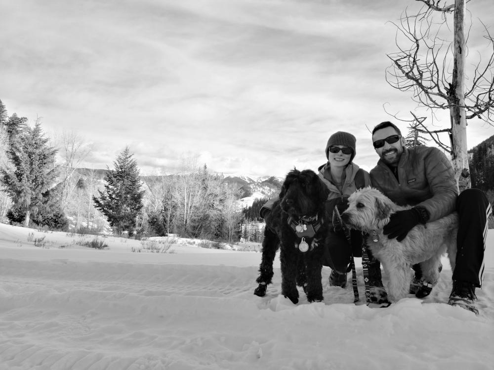 Two people and two dogs kneeling in snow with trees in the background