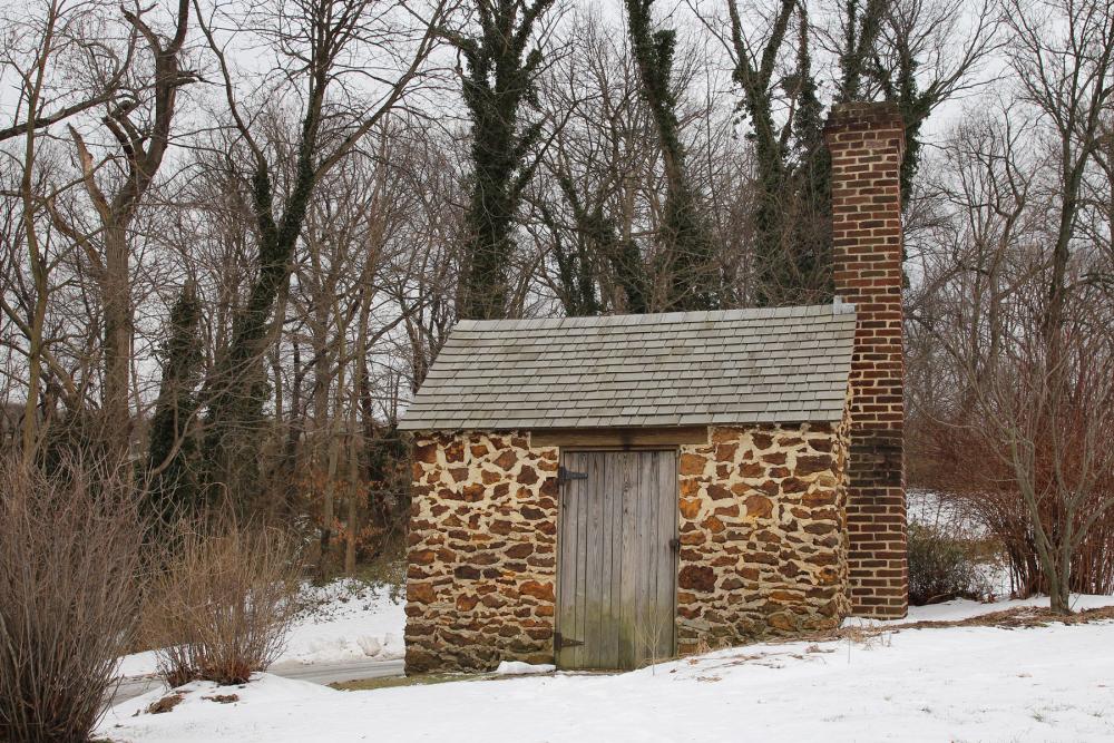 Small windowless brick building with tall brick chimney on snowy ground. Mostly bare trees are visible in midground and background.