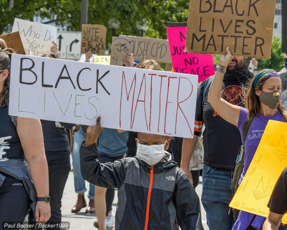 Boy wearing mask and holding sign reading "Black Lives Matter" in front of crowd of protesters 