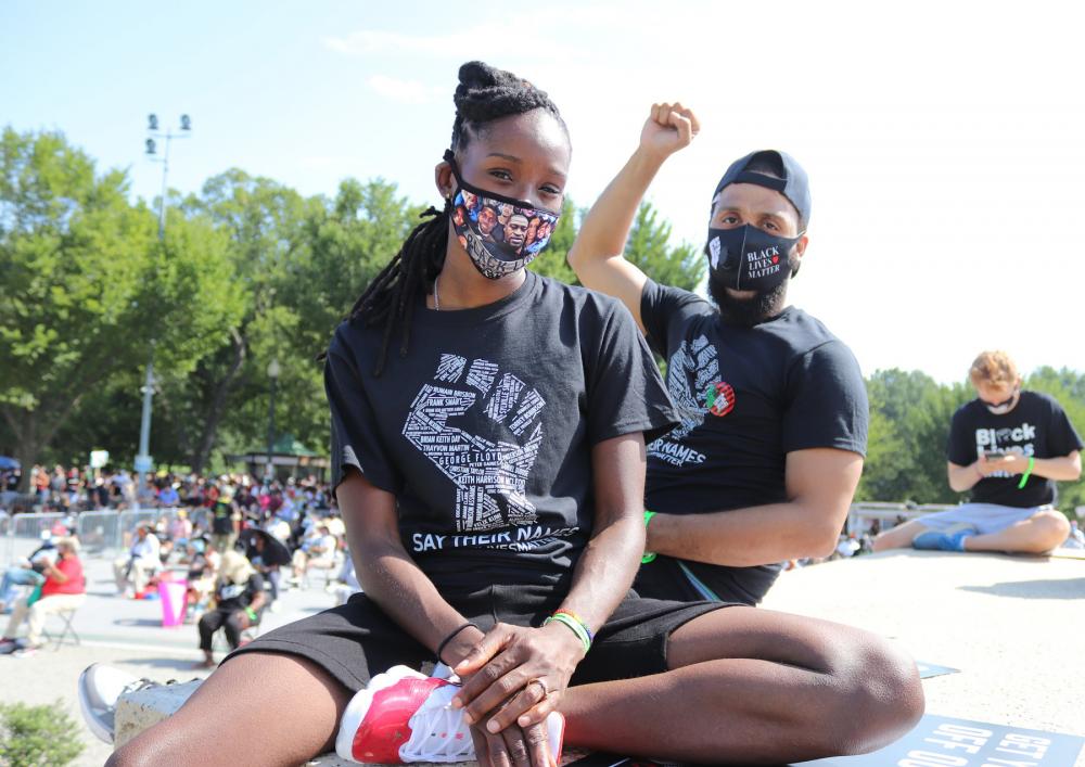 Two protesters seated, wearing masks and looking into the camera, one with a fist raised. Shirts read "Say Their Names" and masks read "Black Lives Matter."