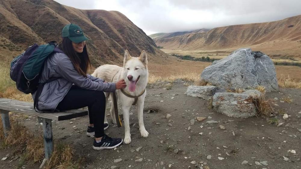 Elizabeth Perez is sitting on a bench petting a white dog. In the background, there is a beautiful mountainous landscape.
