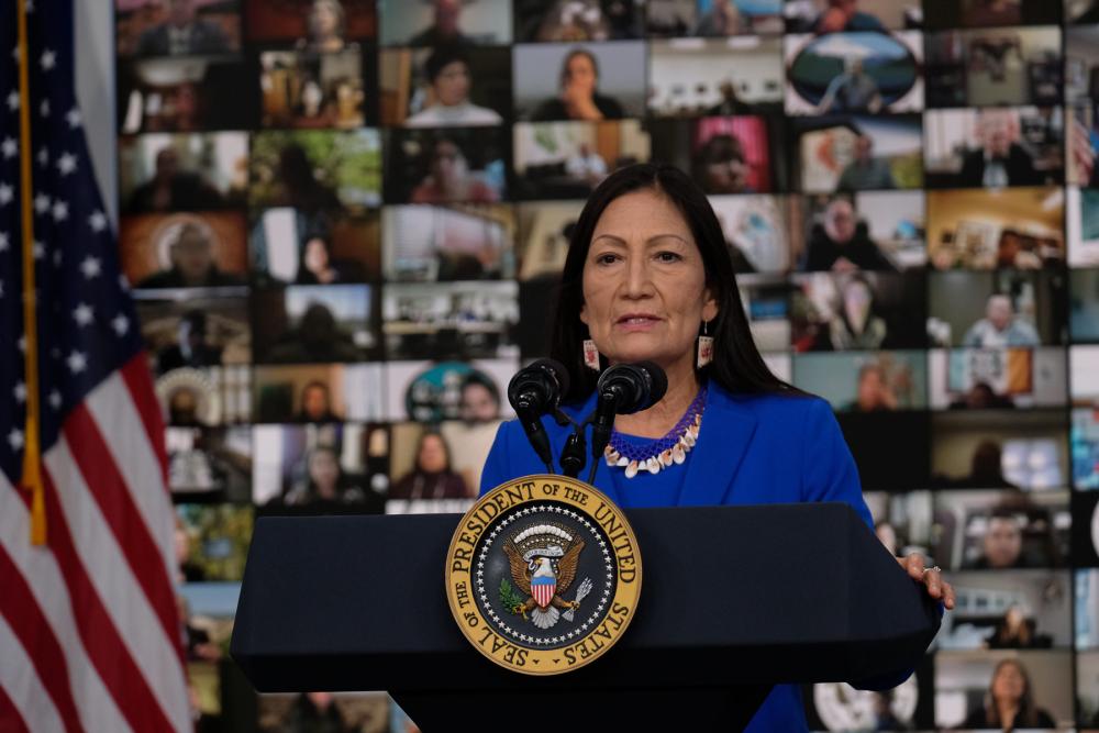 Woman standing at lectern with seal reading "Seal of the President of the United States," speaking into two microphones