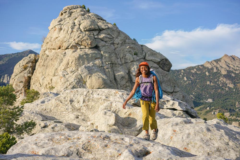 Woman wearing mountain-climbing gear and facing camera, stand-in in front of rocky formation with mountain range in the background