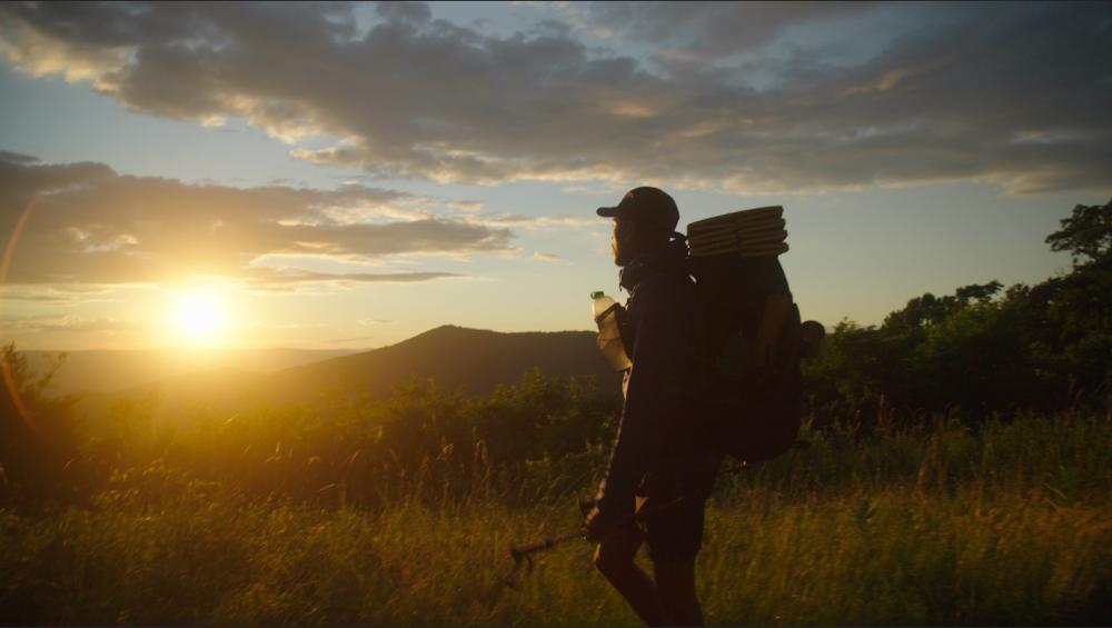 silhouette  of a person carrying a big backpack, with mountains and the sunset in the background