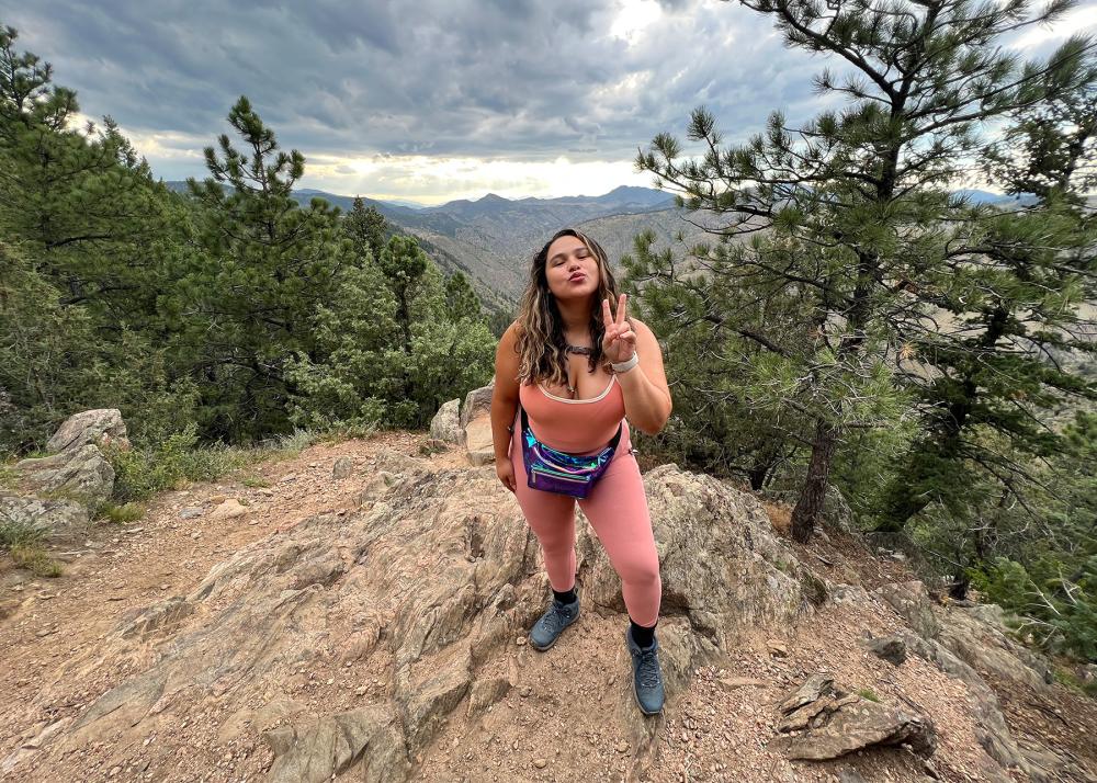 person posing for a picture and doing doing the peace sign, while standing on a hill surrounded by pine trees