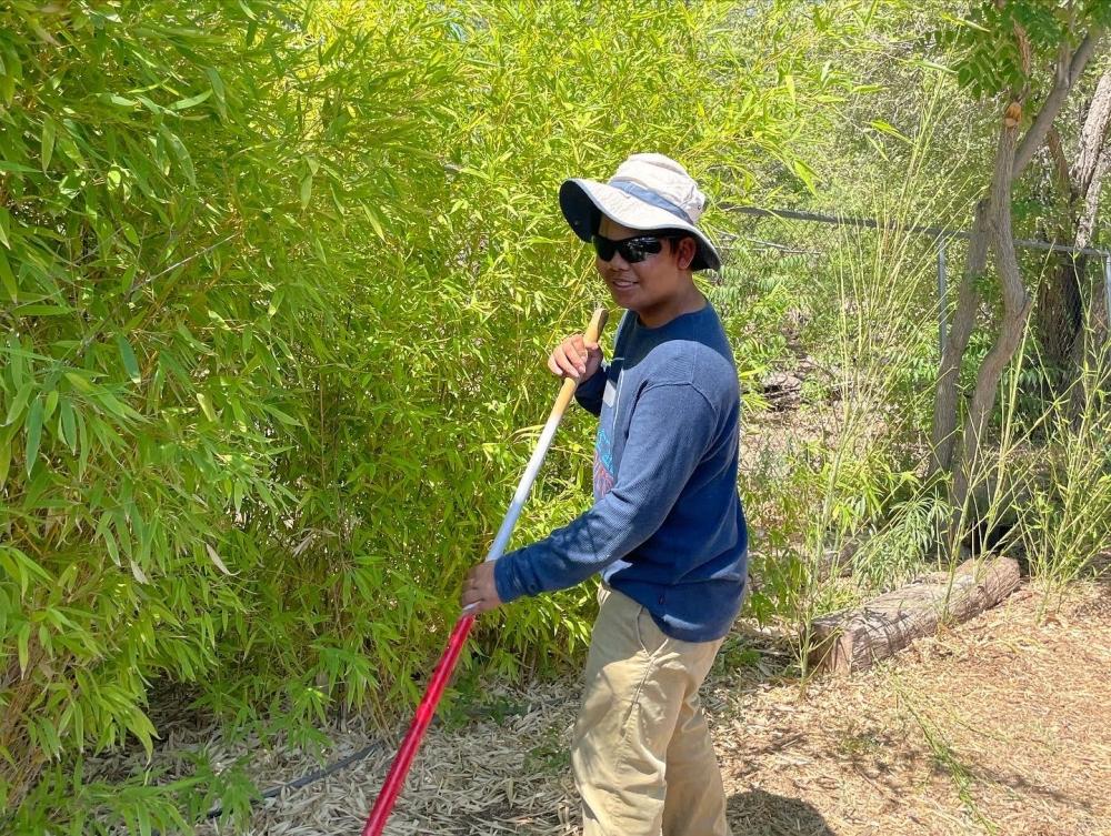 A teenager stands outside holding a gardening tool