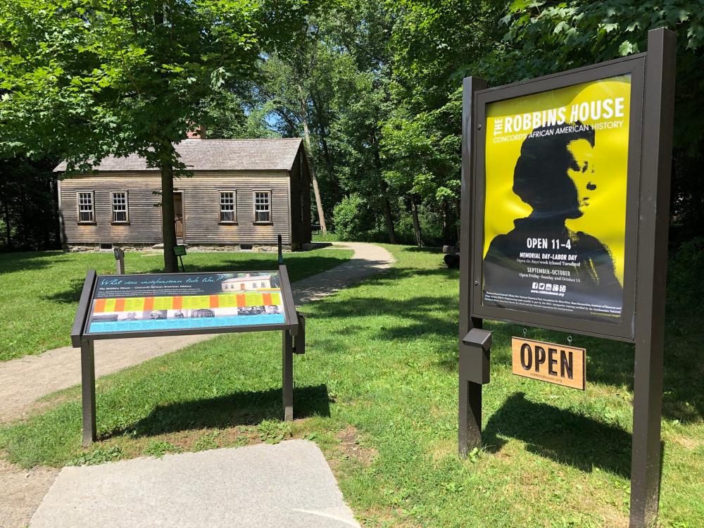 Small house in background amid green trees. Signs in foreground read "The Robbins House" and "Open"