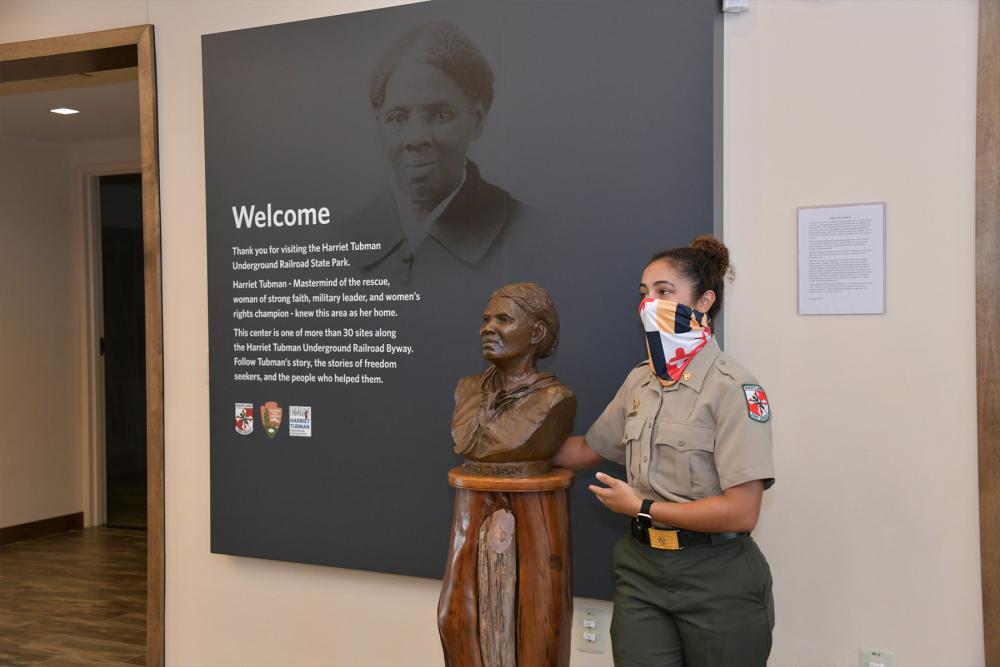 park ranger stands next to bust statue of Harriet Tubman, with a big black and white image of Tubman on the wall behind her